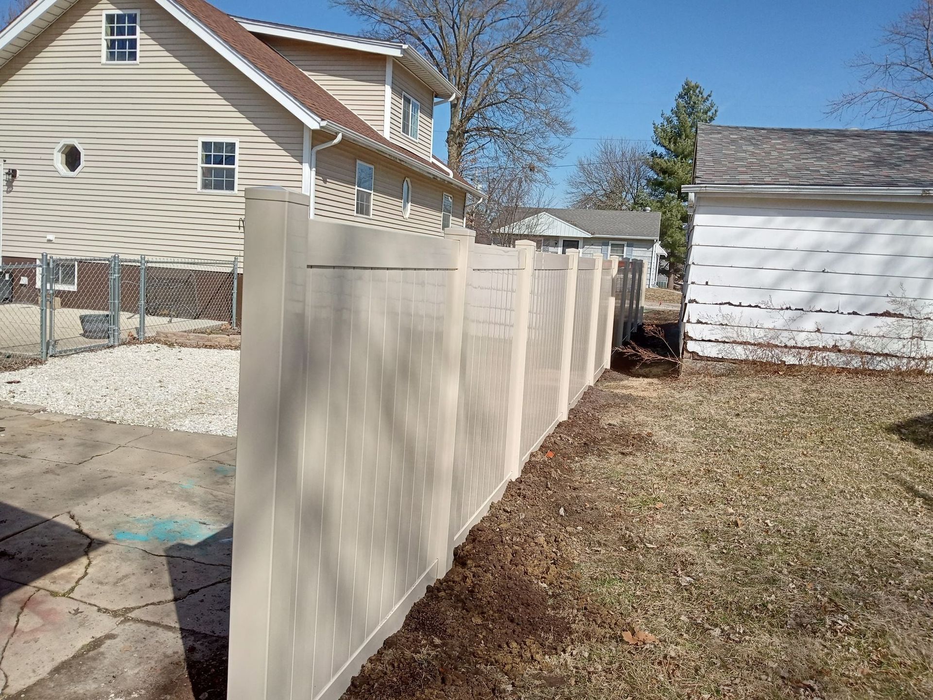 A tan vinyl privacy fence runs alongside a residential backyard, separating a gravel driveway from a grassy yard.