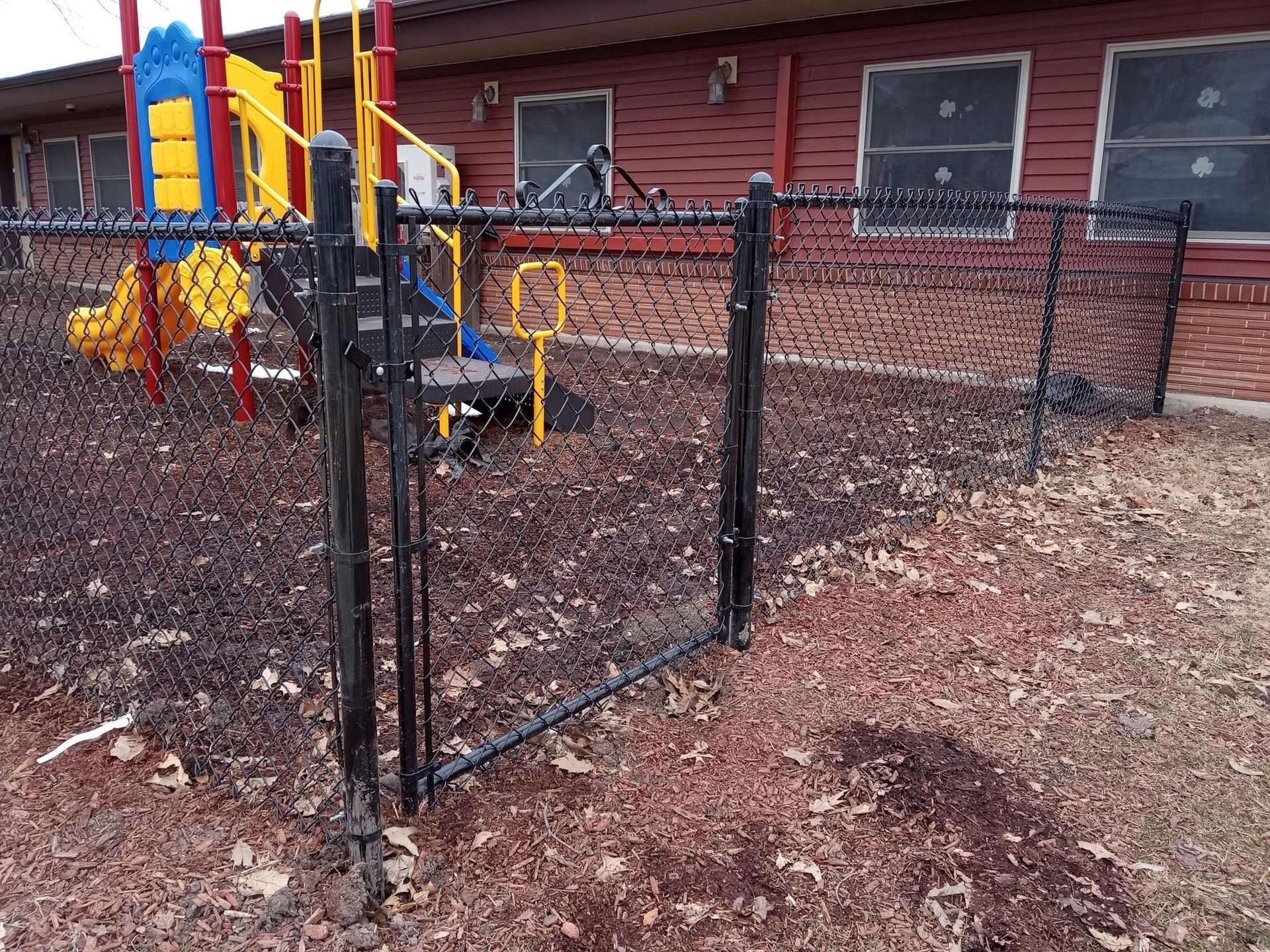 A black chain-link fence surrounds a colorful playground with a slide, set against a red building with windows.