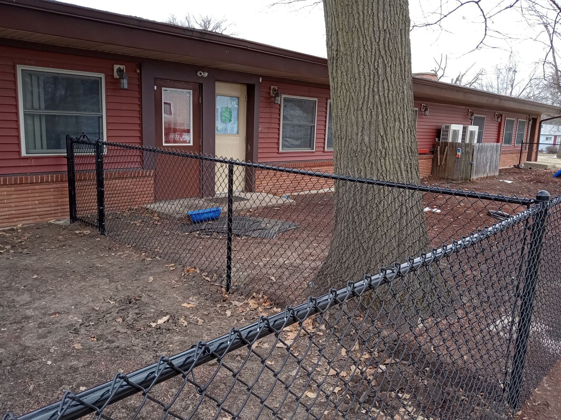 Red building exterior with a tan door, small windows, and a black chain-link fence in the foreground near a tree.