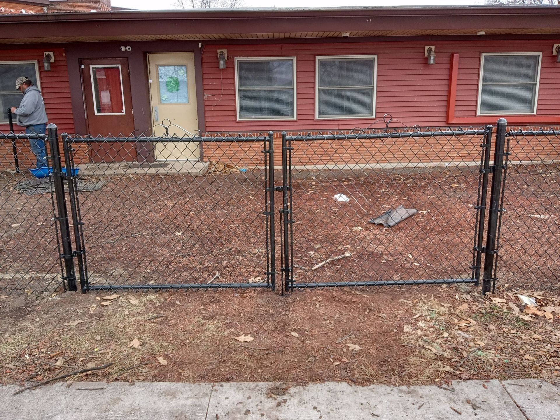 A black chain-link fence stands in front of a red building with several windows and two doors.