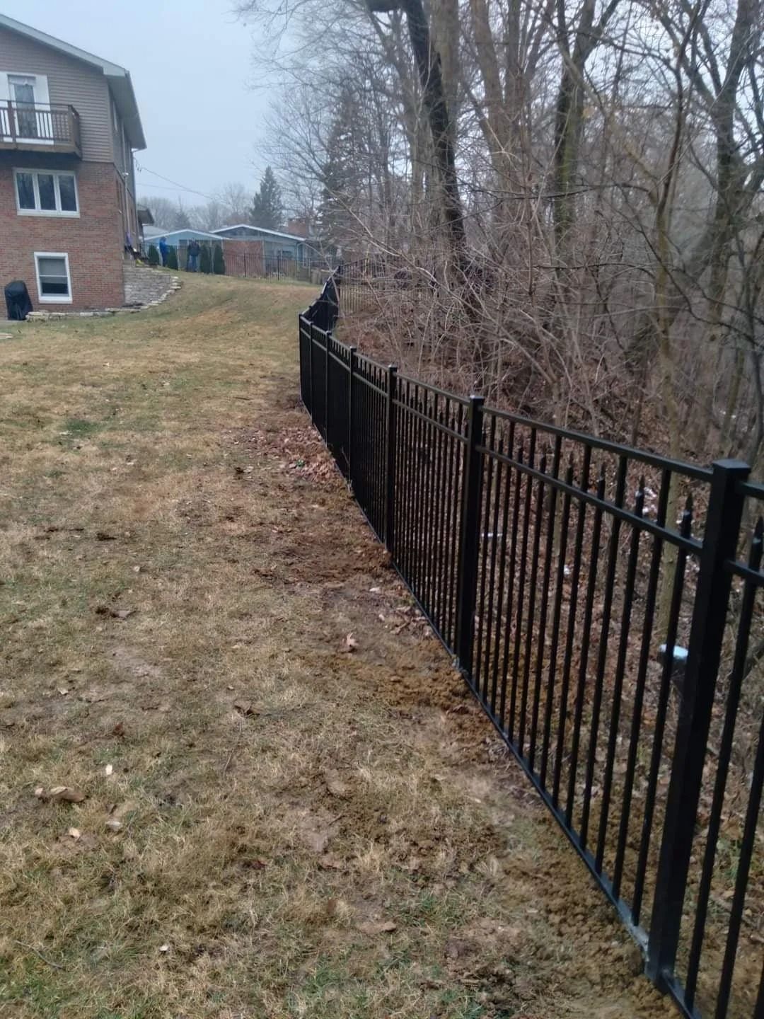 Black metal fence along a grassy slope, next to a building and bare trees.