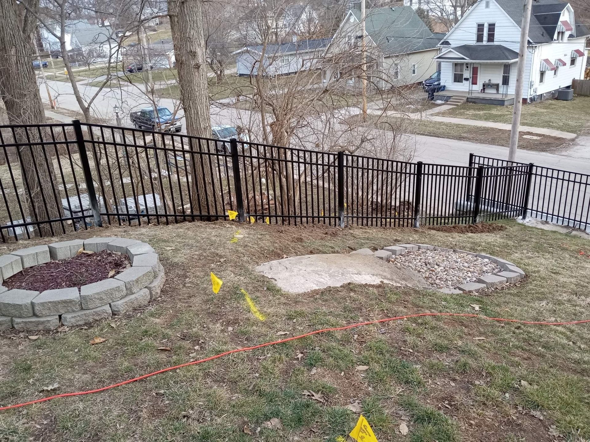 A backyard view featuring a stone fire pit, a concrete patch, a black metal fence, and yellow utility flags on the grass.