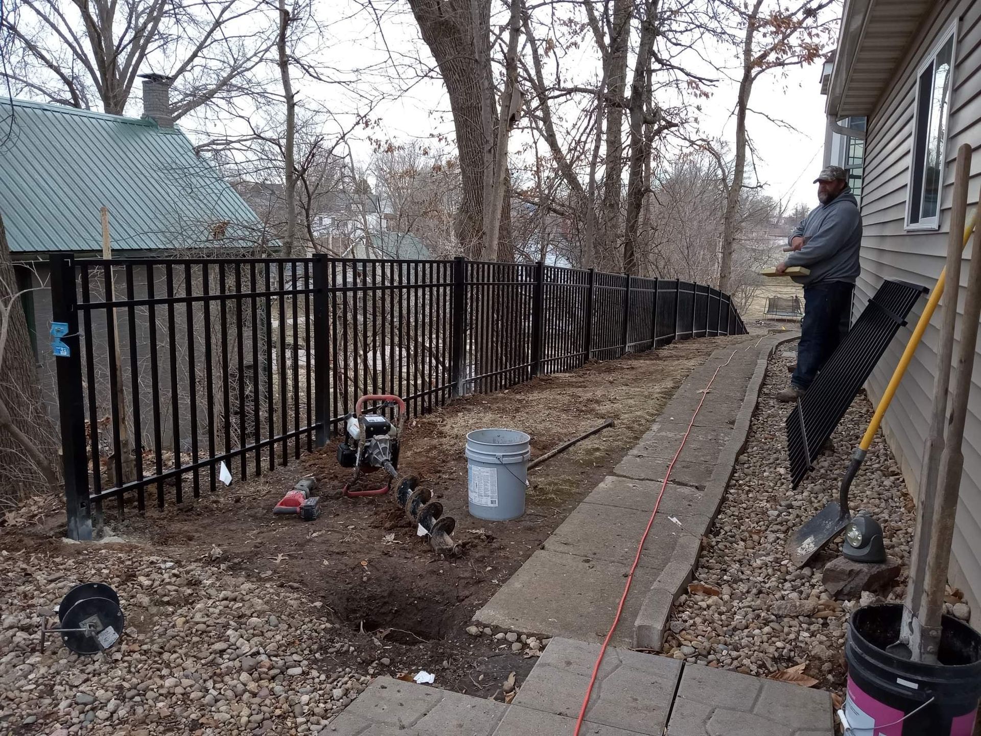 A person stands near a house wall and a newly installed black metal fence, with tools and a post hole on the ground nearby.