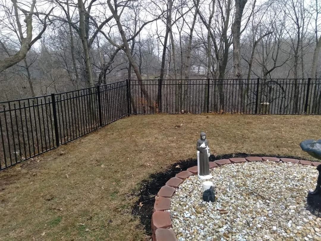 Black metal fence on a hillside, surrounding a brown lawn area. Bare trees in the background.
