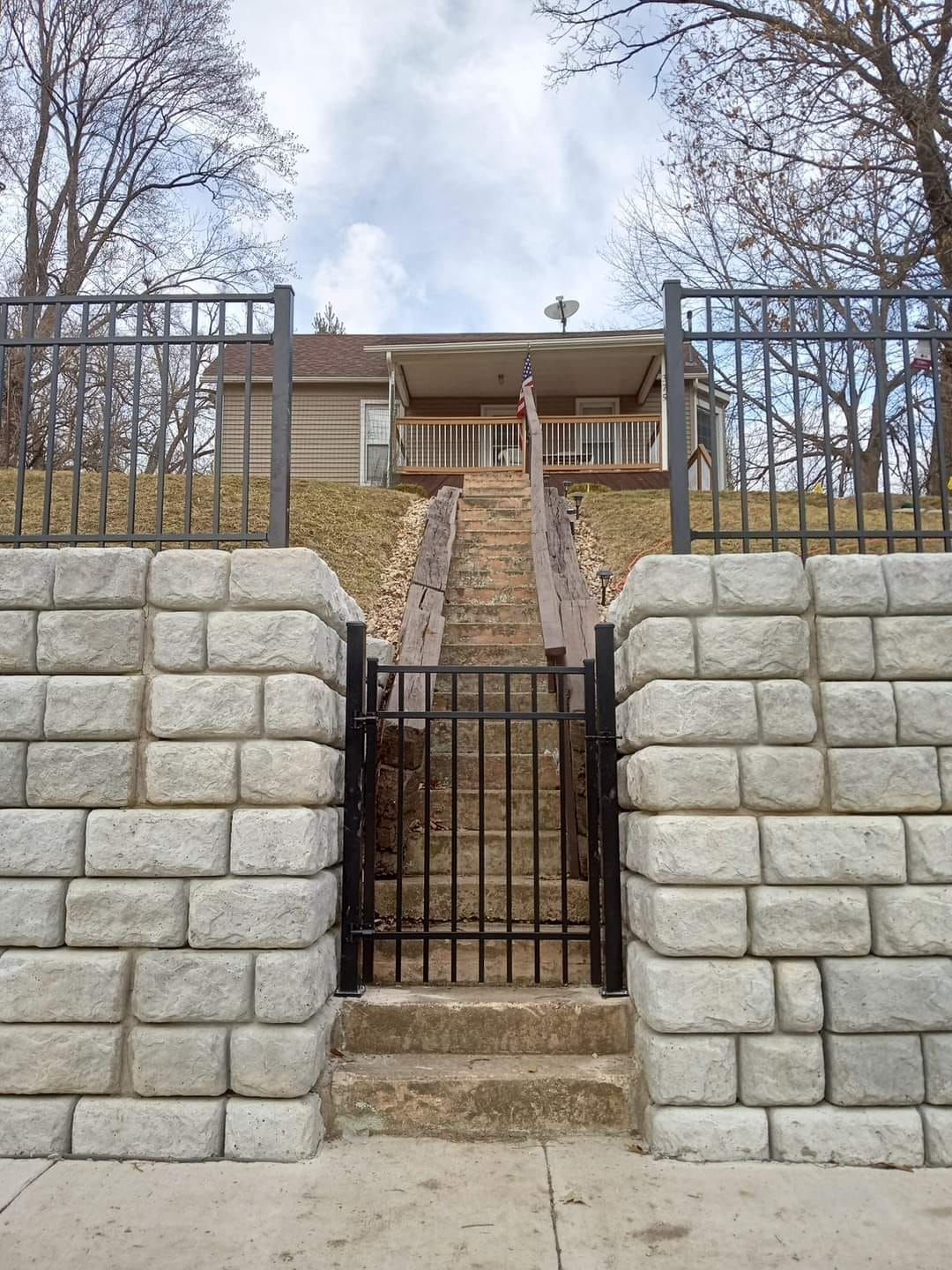 A stone retaining wall with a black metal gate leads to a set of stairs rising toward a house on a grassy hill.