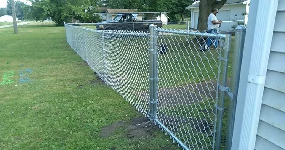 Chain-link fence in a grassy yard, with a gate near a white house. A person stands nearby.