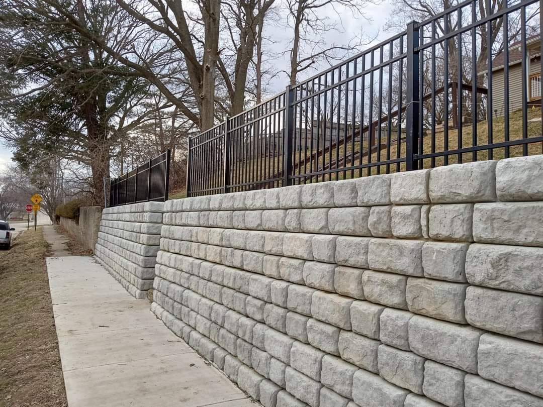 A tall, light gray concrete block retaining wall lines a public sidewalk, topped with a black metal safety fence.