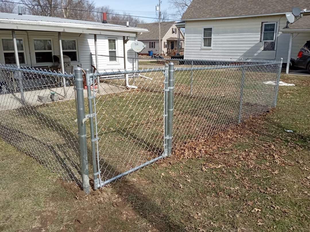 A chain-link fence gate standing open in a backyard, with a white house in the background.
