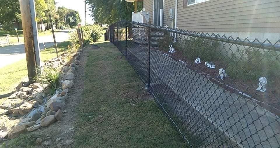 Black chain-link fence along a grassy strip next to a building and stone retaining wall.