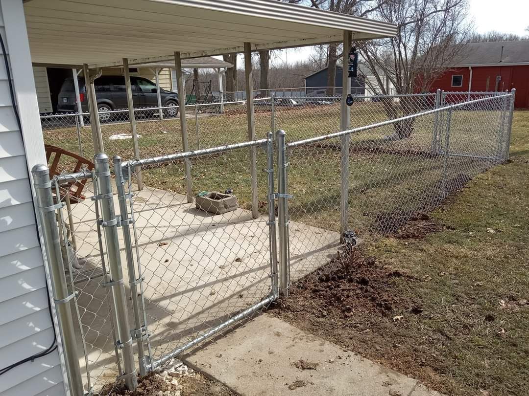 A chain-link fence and gate installed on a concrete patio next to a house, with a yard and shed in the background.