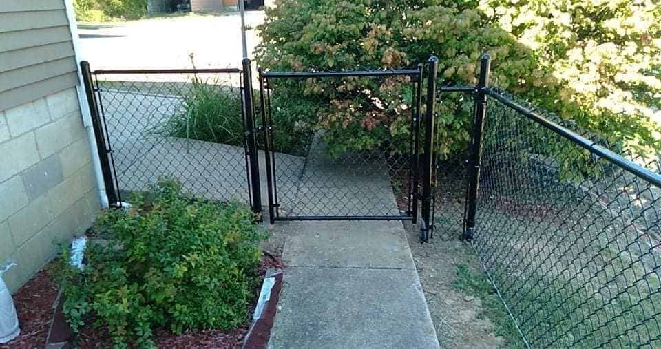 Black chain-link fence with gate blocks a concrete path. Lush bushes flank the fence.