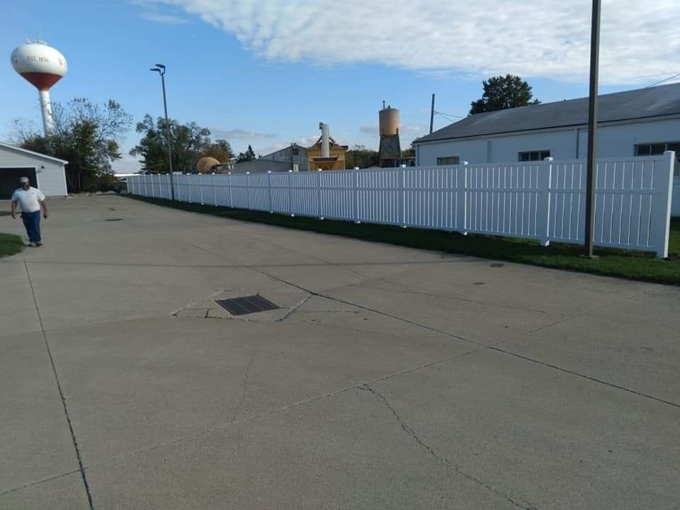 A person walks on a concrete lot past a long white vinyl fence toward industrial buildings and a water tower.