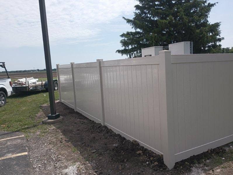 Beige vinyl fence along a sidewalk, with a grassy lot and parked vehicles nearby under a cloudy sky.