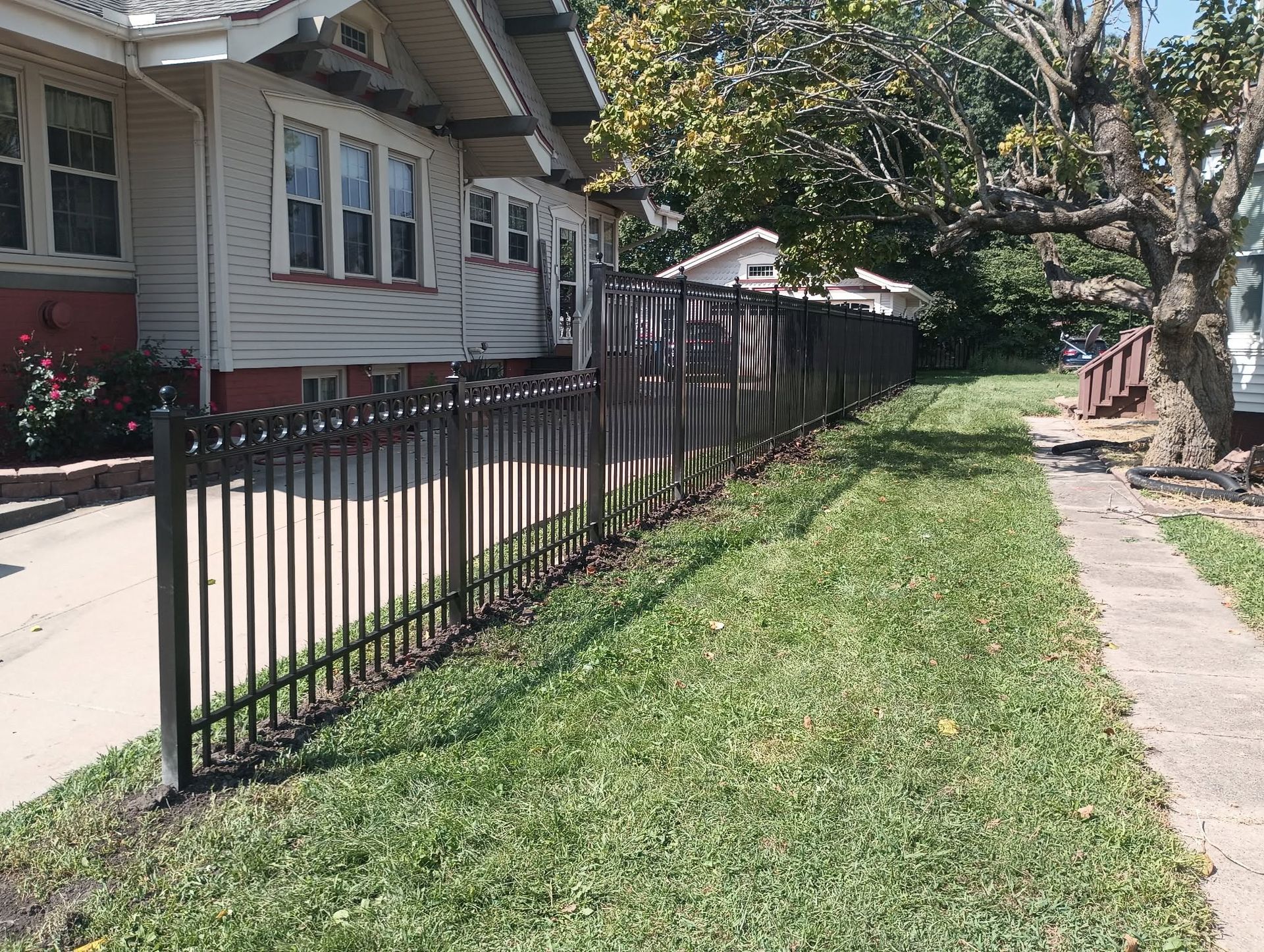 Residential side yard with a black metal fence, sidewalk, and house along a grassy strip