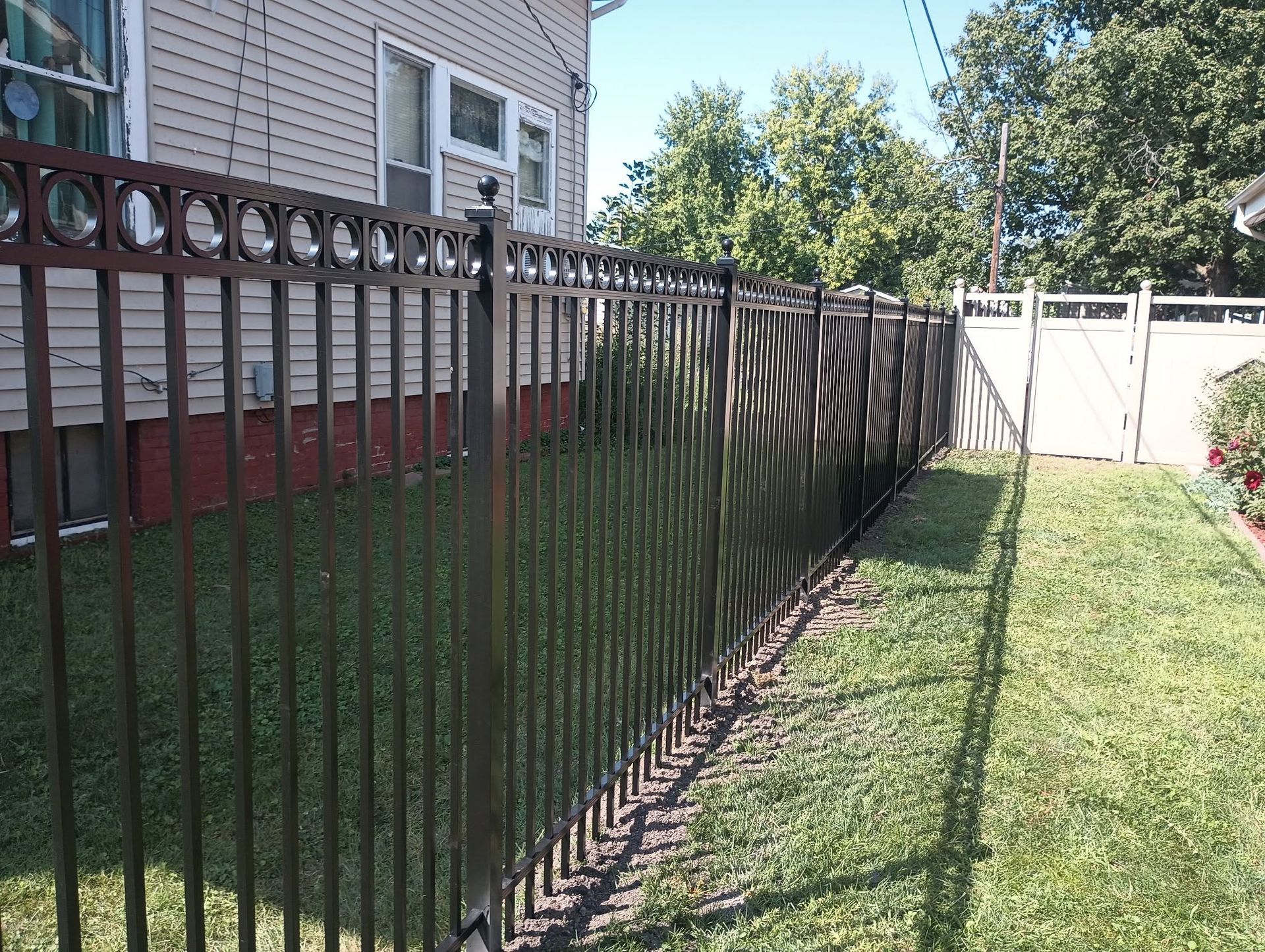 Black metal fence along a grassy side yard beside a house, with trees and a white fence in the background