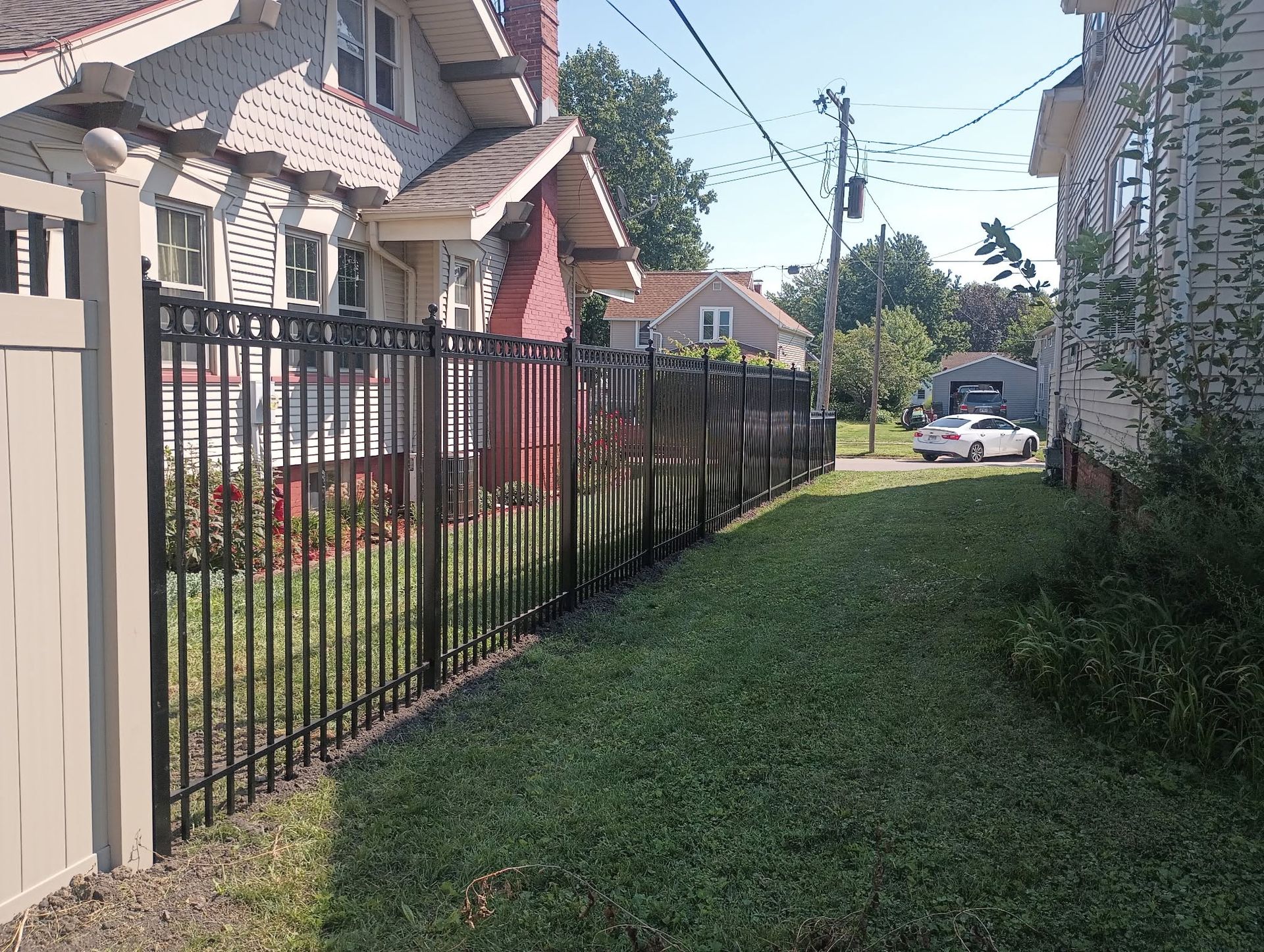 Narrow grassy alley beside houses and black iron fence, leading to a street with a parked car.