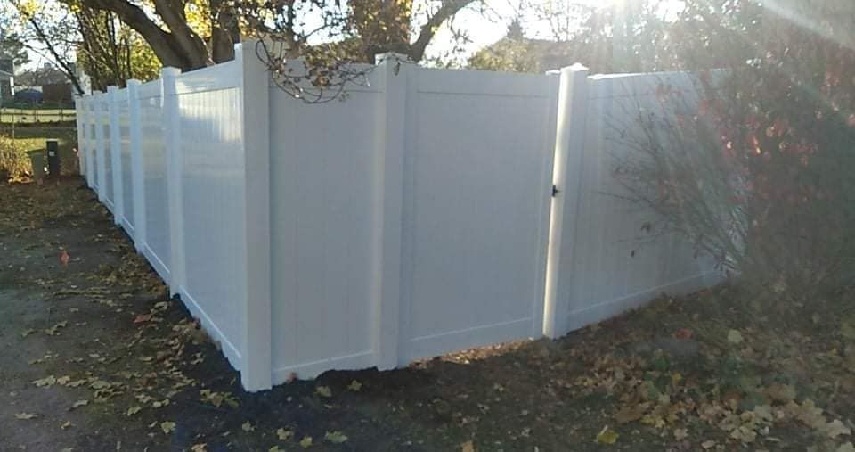 A white vinyl privacy fence with a closed gate stands on a lawn covered in fallen autumn leaves.