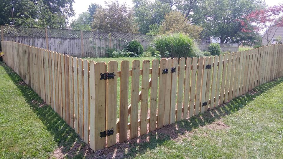 A light-wood picket fence with a gate stands in a grassy yard, with an older wooden fence and trees in the background.