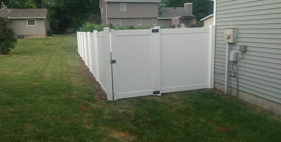 A white vinyl privacy fence with a closed gate extends from the side of a house into a grassy residential backyard.