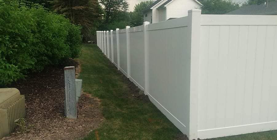A long white vinyl privacy fence runs along a green lawn beside a line of bushes.