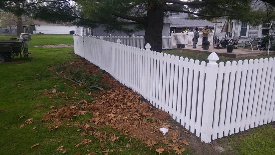 A white picket fence runs along a yard covered in fallen leaves, with a tree and a house patio visible in the background.