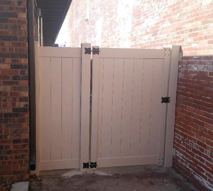 A double-gate off-white vinyl fence stands between two brick walls in an outdoor setting.