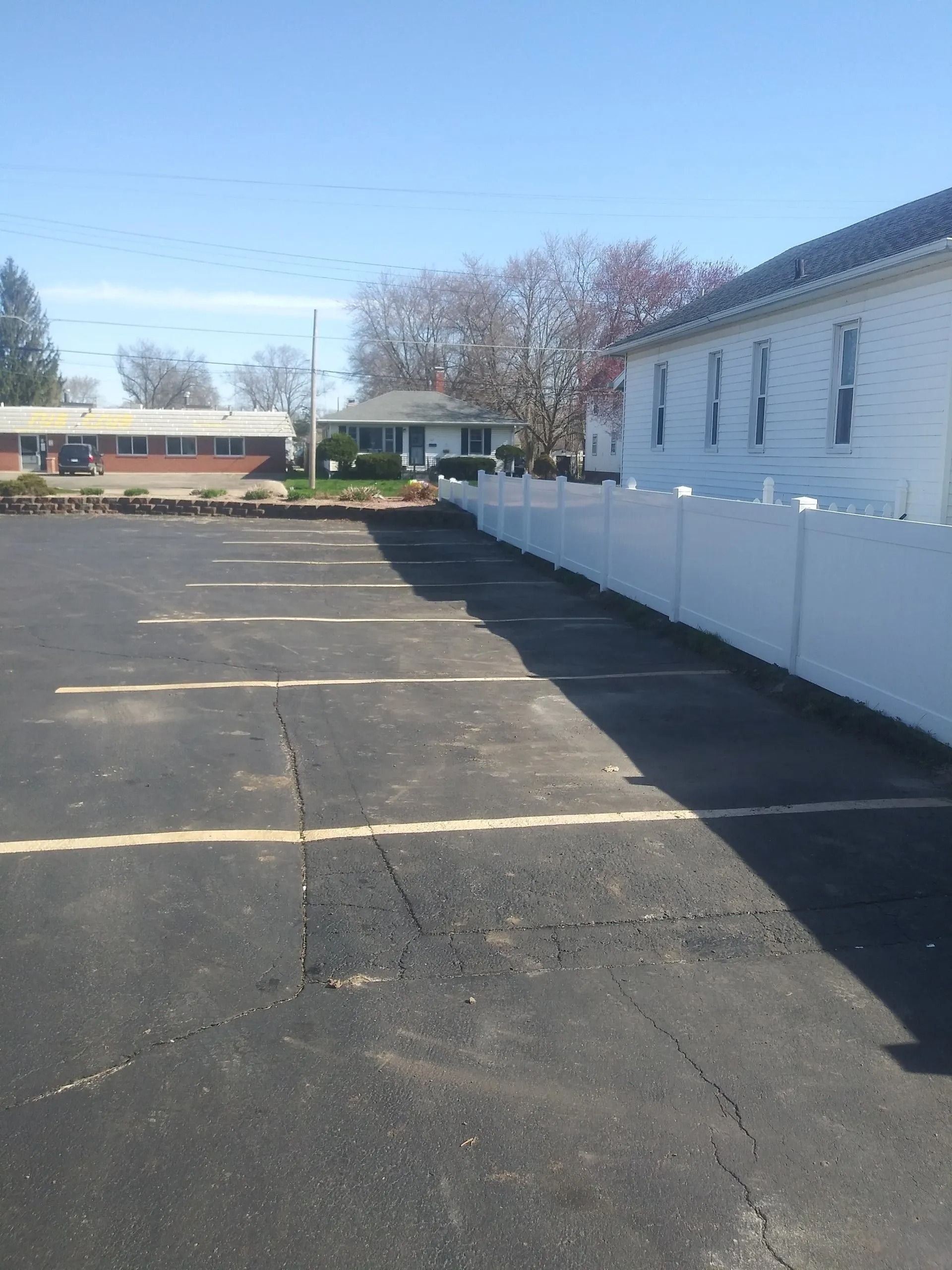 Asphalt parking lot with white fence, building, and trees against a blue sky.