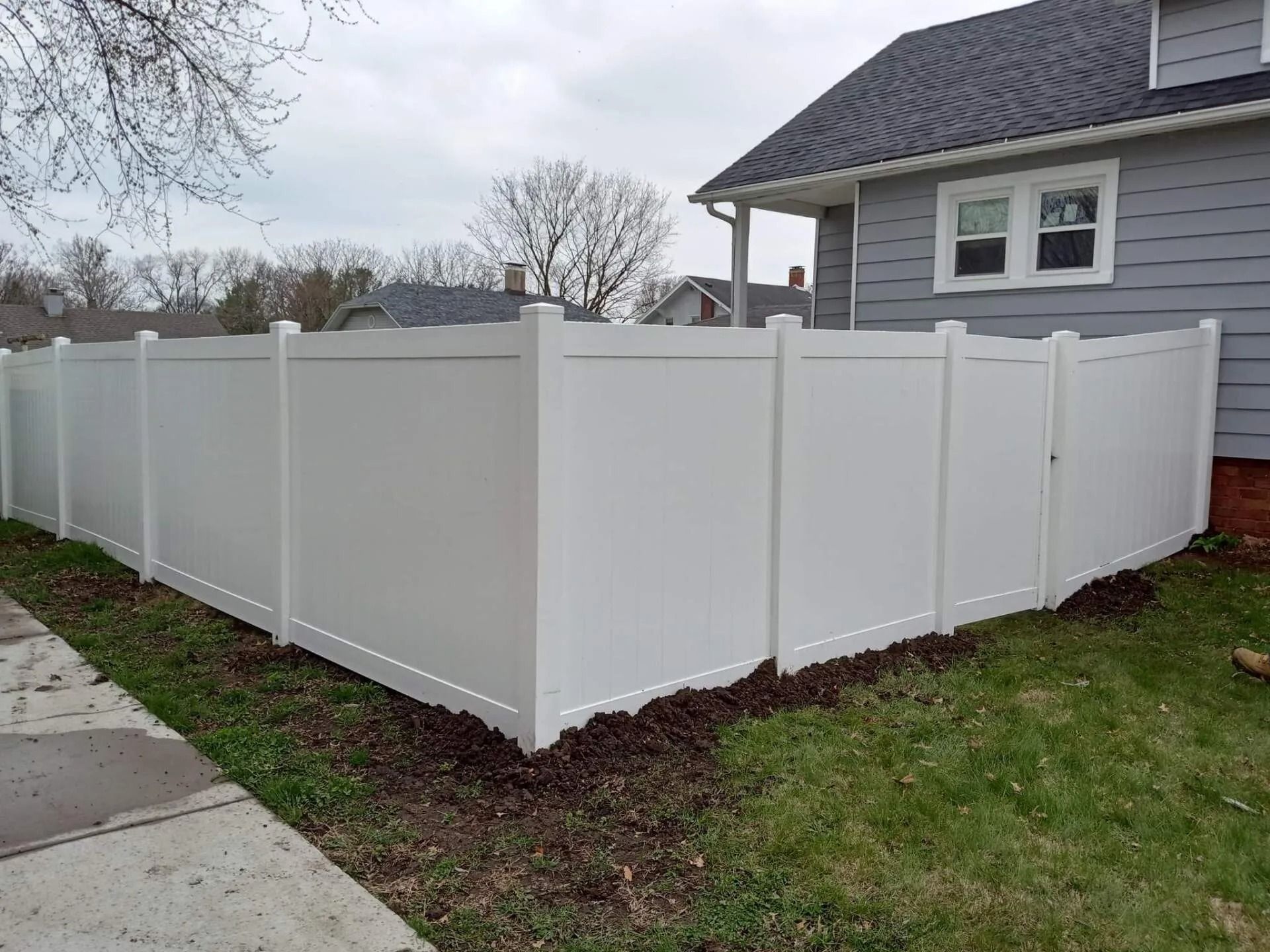 White vinyl fence surrounds a corner lot next to a gray house; cloudy sky.