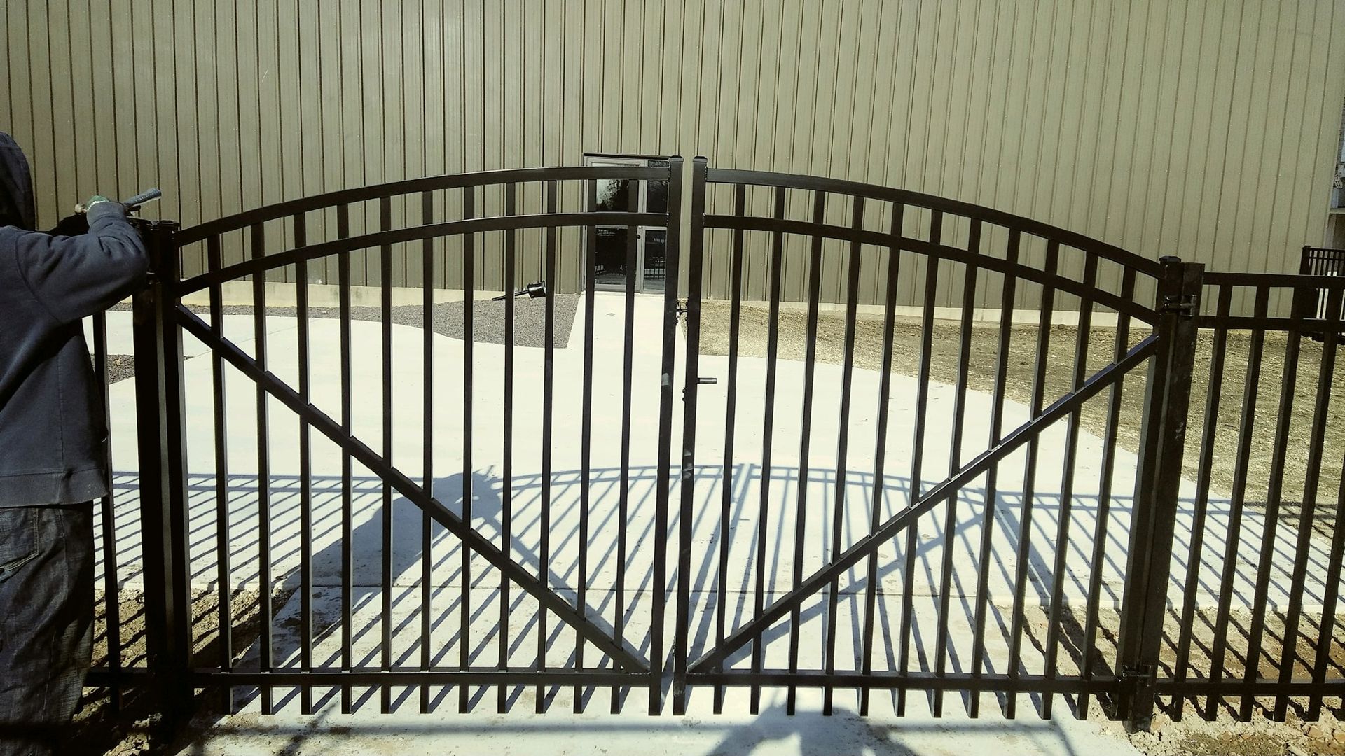A person stands next to a black, arched metal double gate installed in front of a tan, vertically ribbed building.