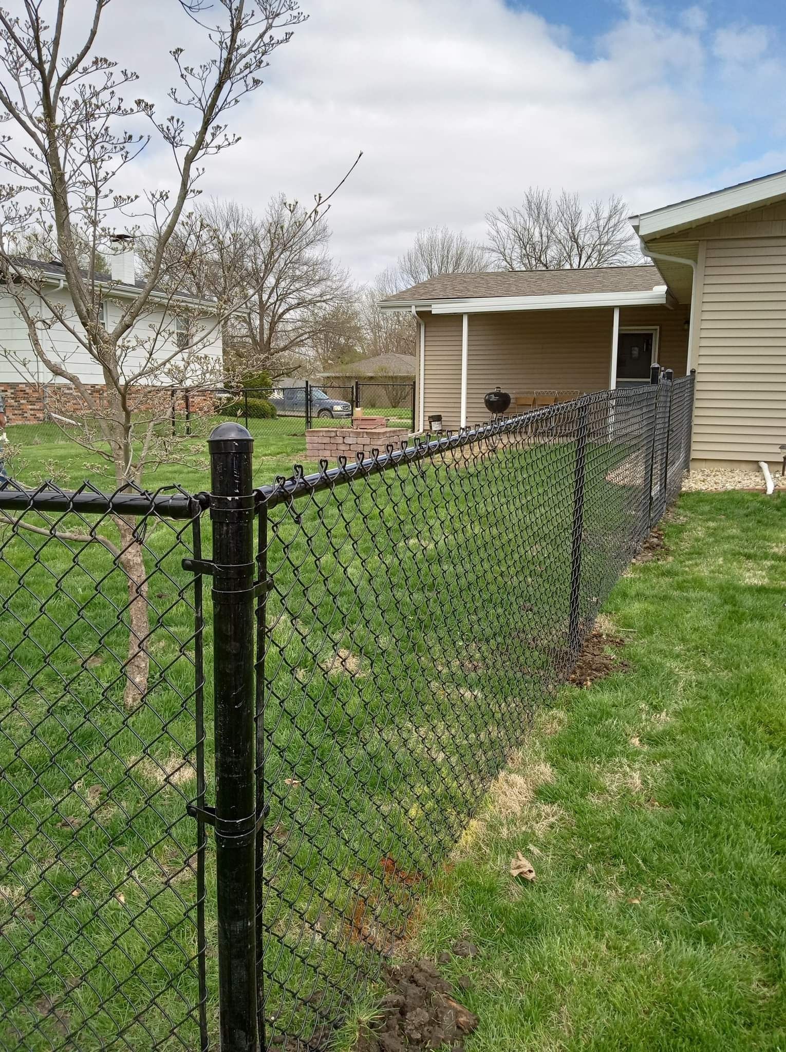 Black chain-link fence with privacy slats bordering a grassy yard, next to a beige house, under a cloudy sky.