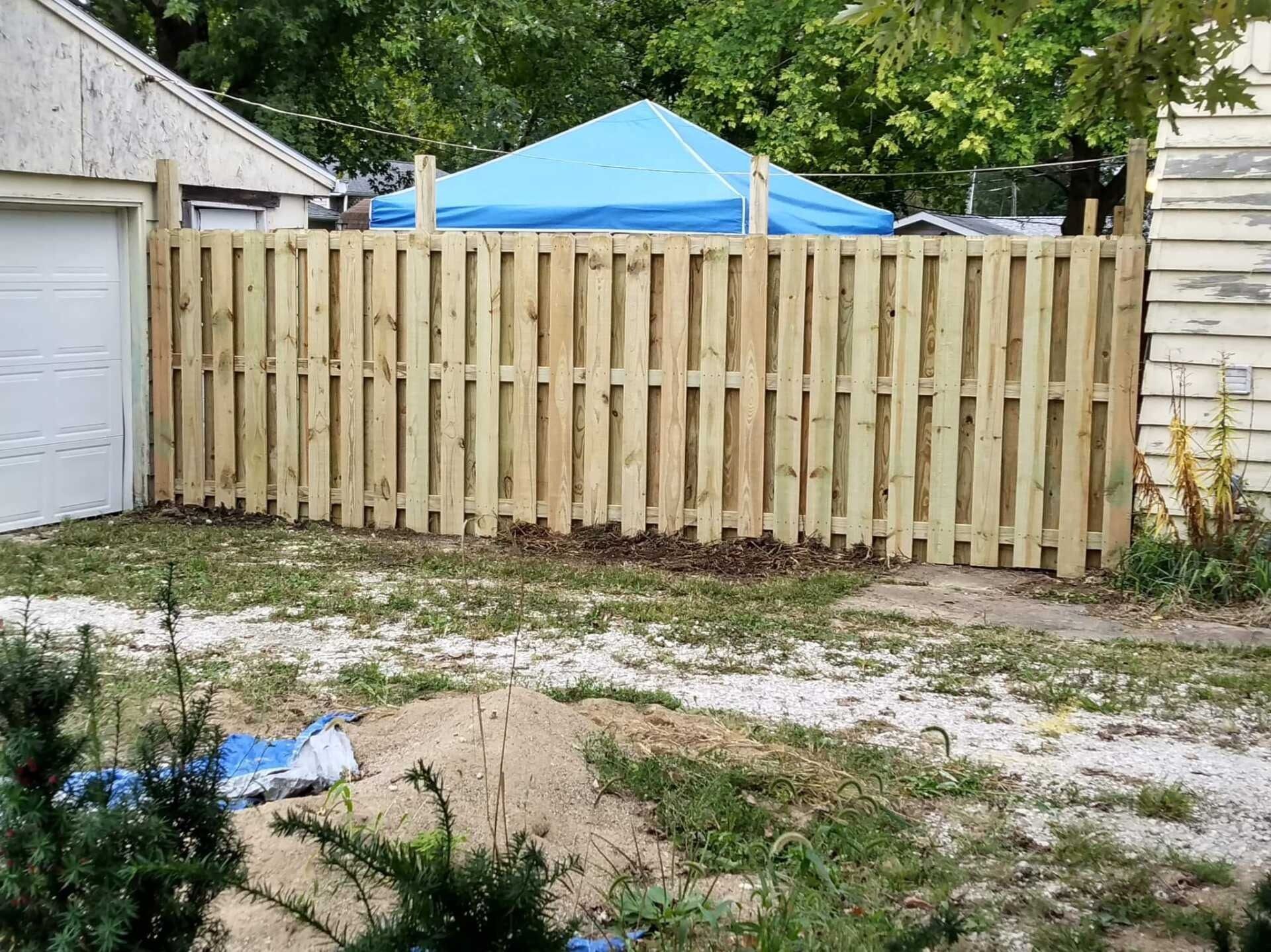 Wooden fence in a yard, near a garage and overgrown grass. A blue canopy is in the background.