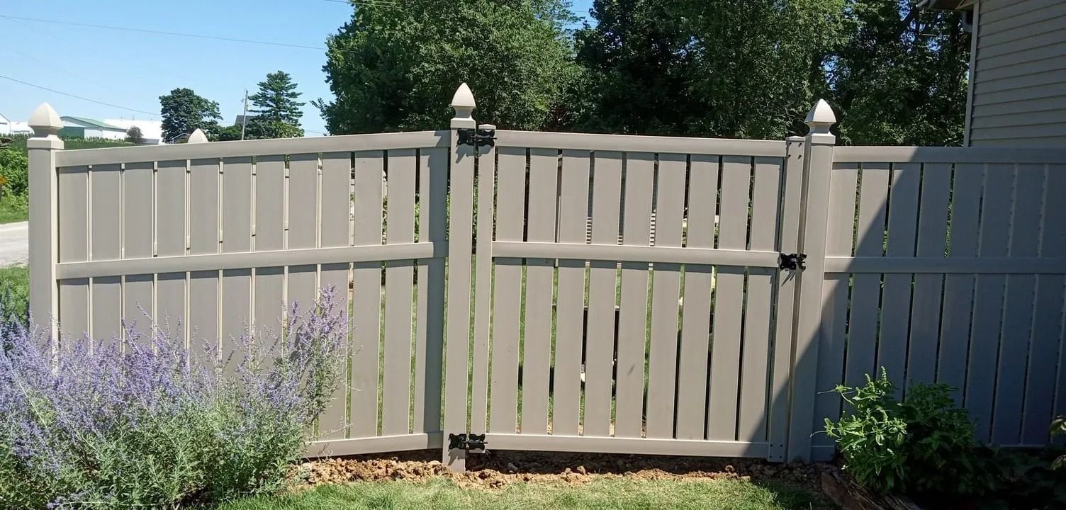 A gray wooden fence with a gate, topped with decorative finials, in a yard with greenery and a blue sky.