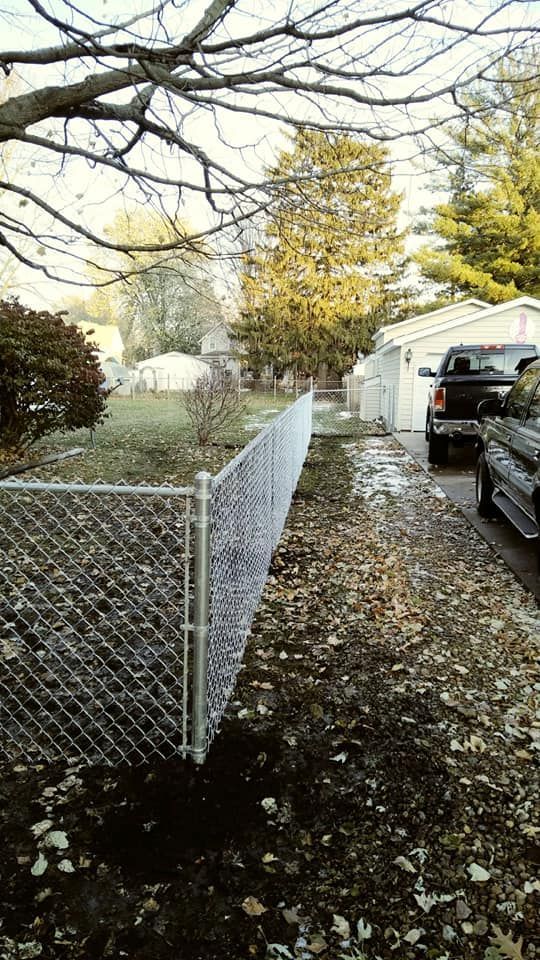 A chain-link fence lines a yard covered in fallen leaves next to a driveway with a parked truck on a cloudy day.