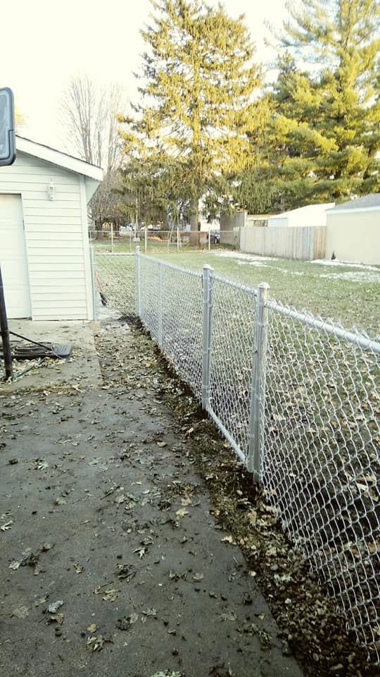 A chain-link fence runs alongside a paved driveway near a white building with trees in the background.