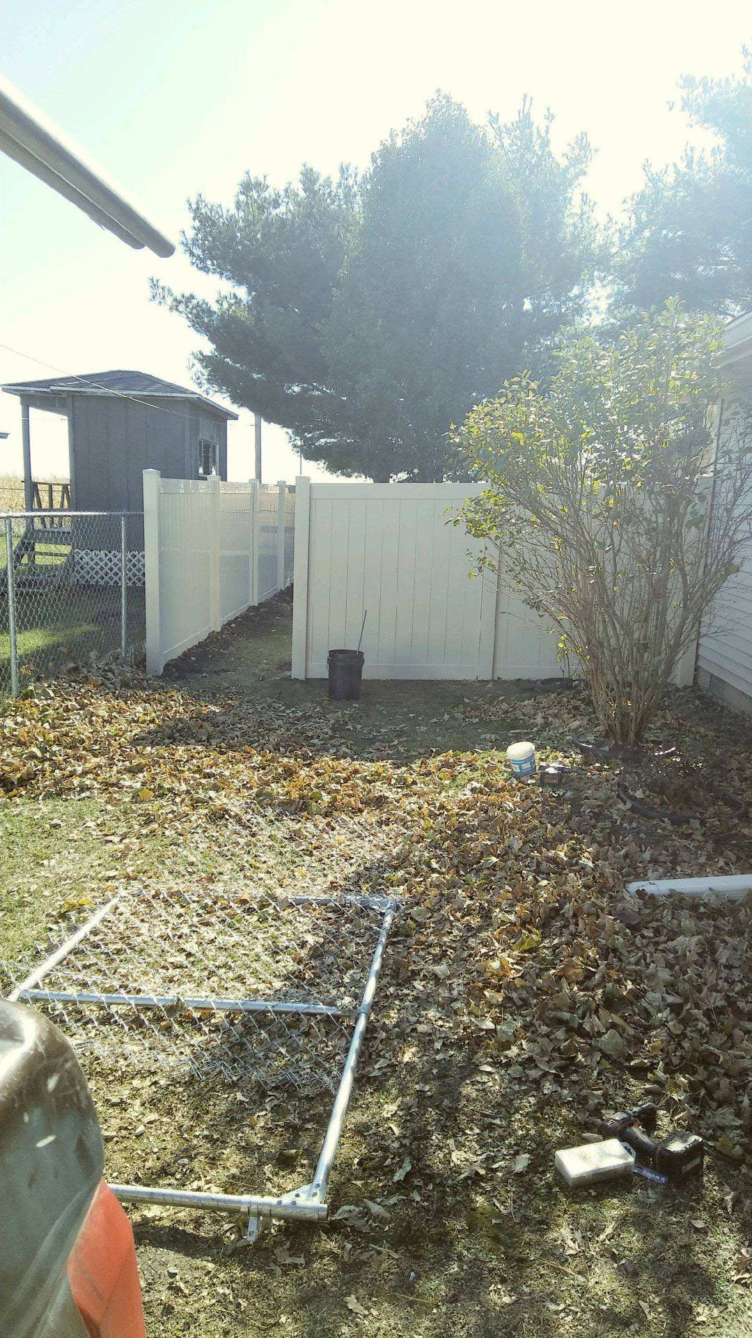A residential backyard scene with scattered leaves, a white privacy fence, a small shed, a shrub, and a metal frame.