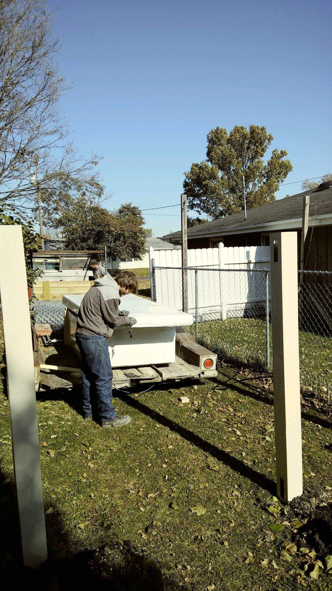 A person standing by a flatbed trailer in a grassy yard, positioned between two vertical wooden posts.