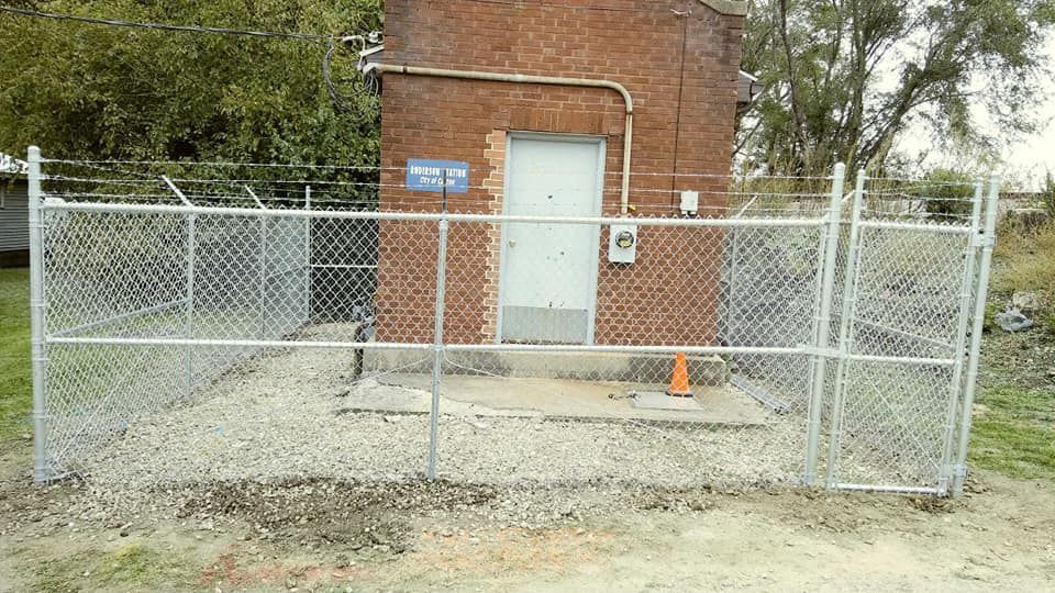A brick utility building behind a chain-link fence, with a white door, a blue sign, and an orange traffic cone.