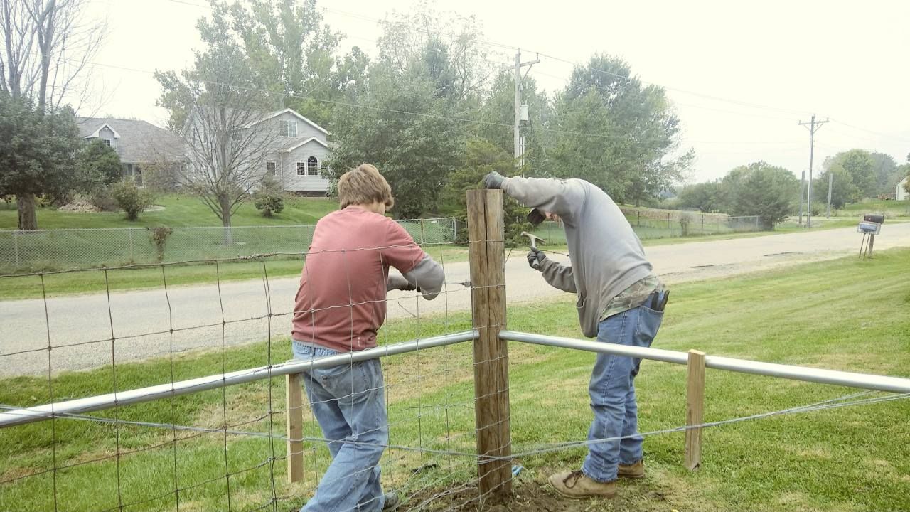 Two people work together to secure a horizontal metal rail to a wooden fence post in a grassy, suburban outdoor setting.