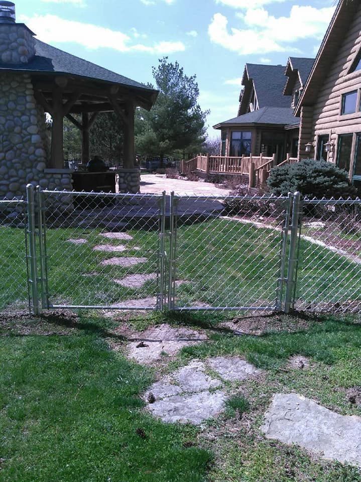 A chain-link fence gate on a stone path leads to a wooden patio and a log cabin in a grassy backyard.