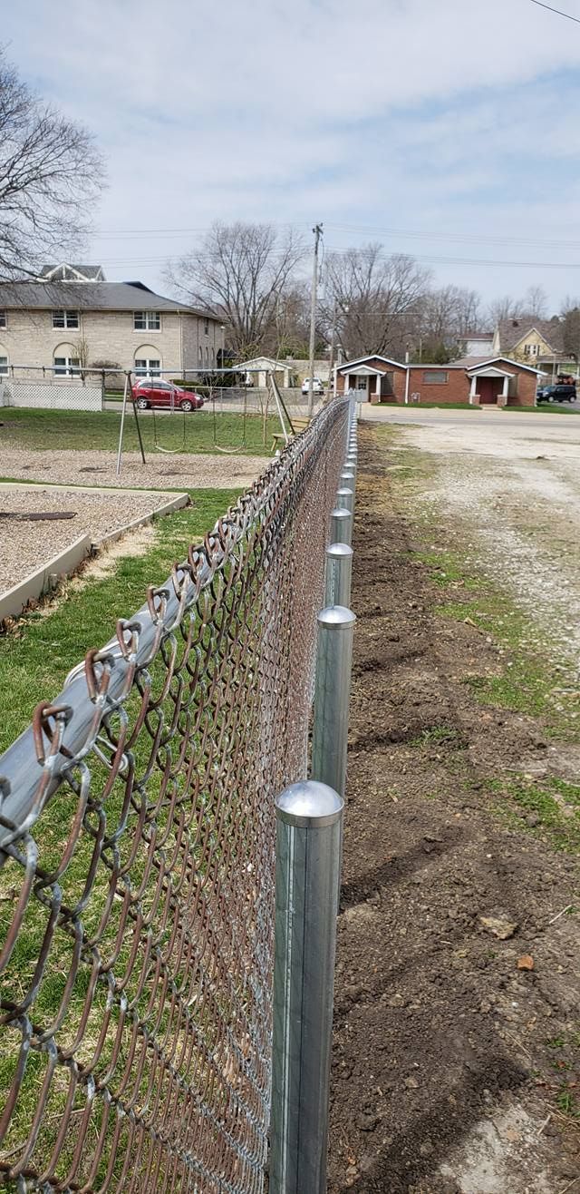 A chain-link fence with silver metal posts stands along a dirt path bordering a residential area.