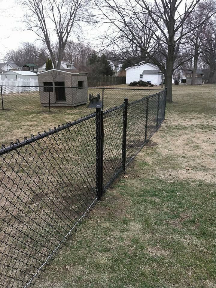 A black chain-link fence runs through a grassy backyard with a small playhouse in the background under an overcast sky.