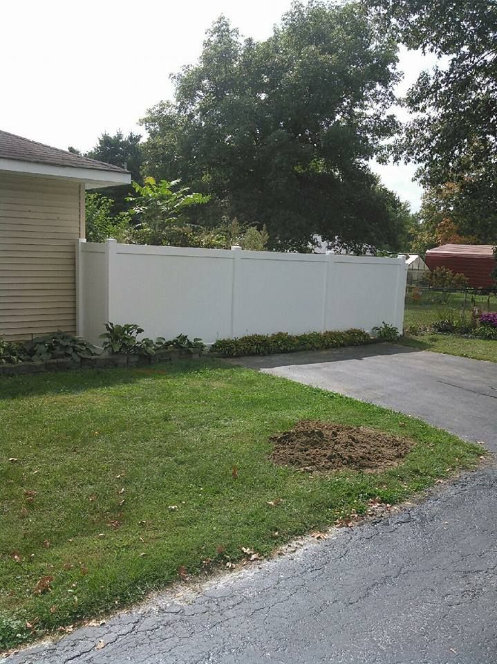 A white privacy fence runs along a paved driveway next to a tan house and green lawn on a sunny day.