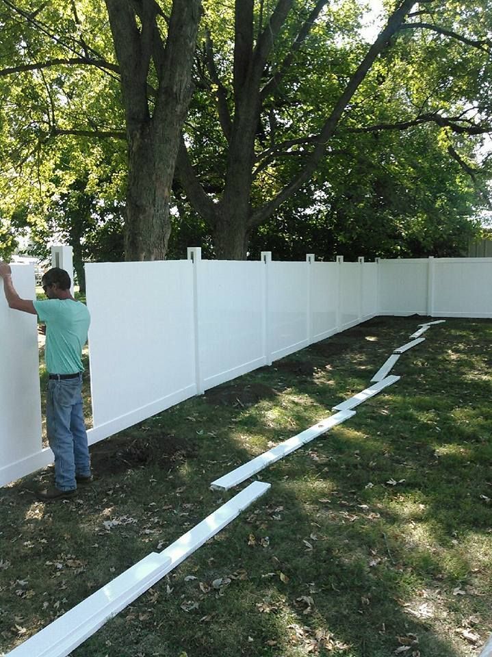 A person in a light green shirt installing white vinyl fencing panels in a backyard next to a large tree.