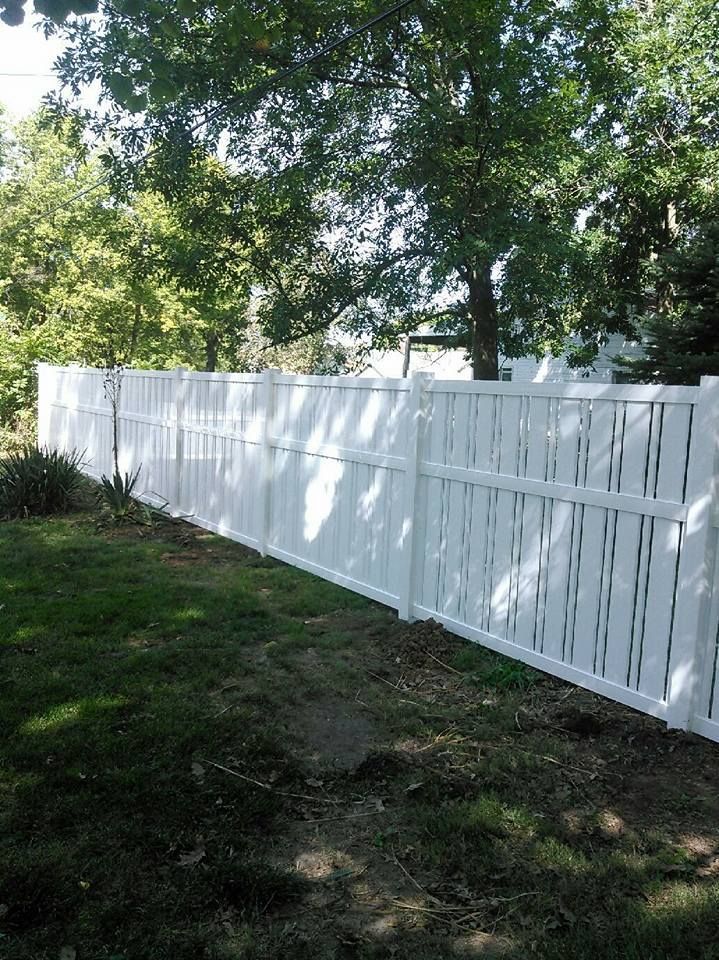 A white vinyl privacy fence stands in a grassy yard, partially shaded by green trees.