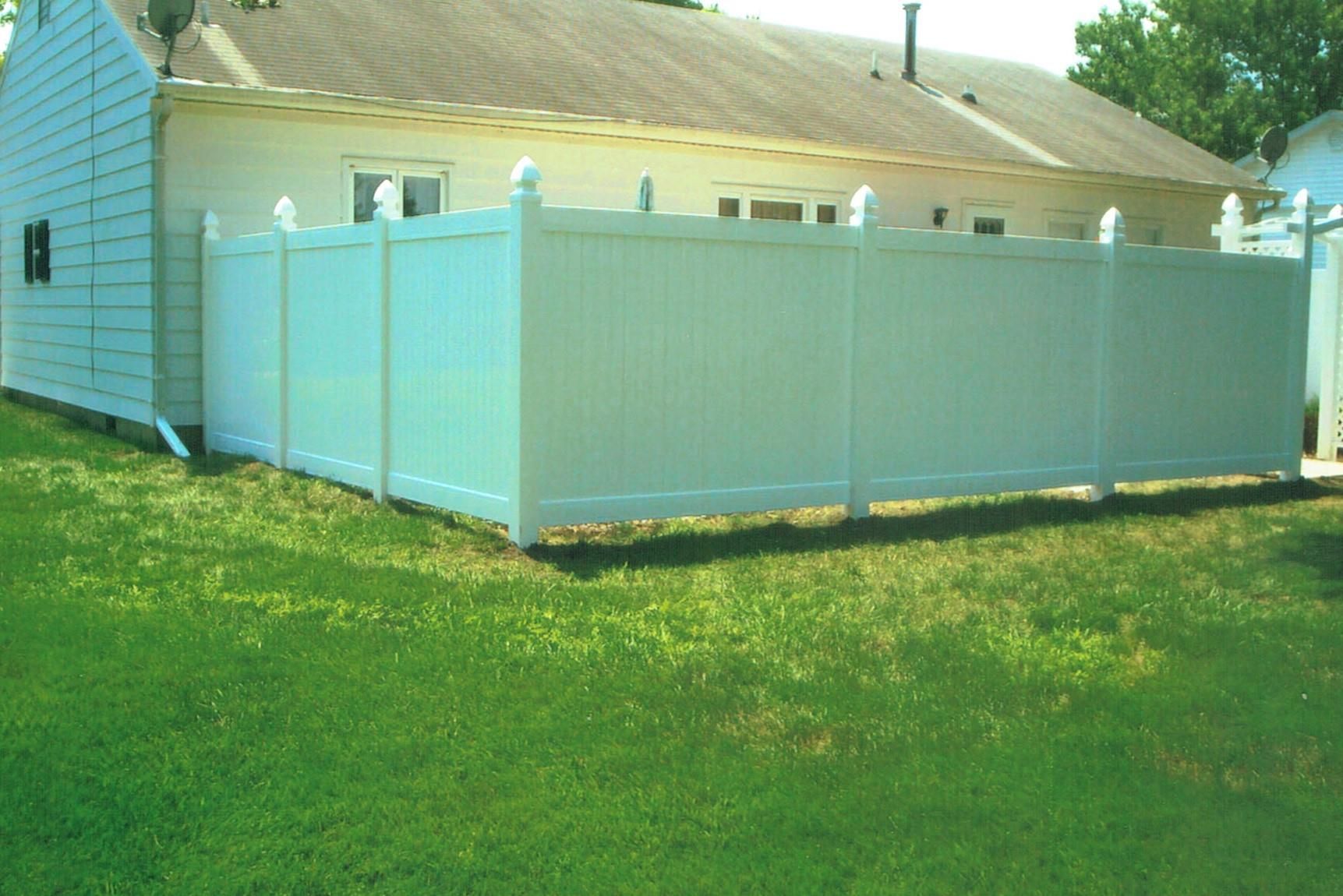 A white vinyl privacy fence encloses the side and back of a house with light blue siding and a shingled roof.