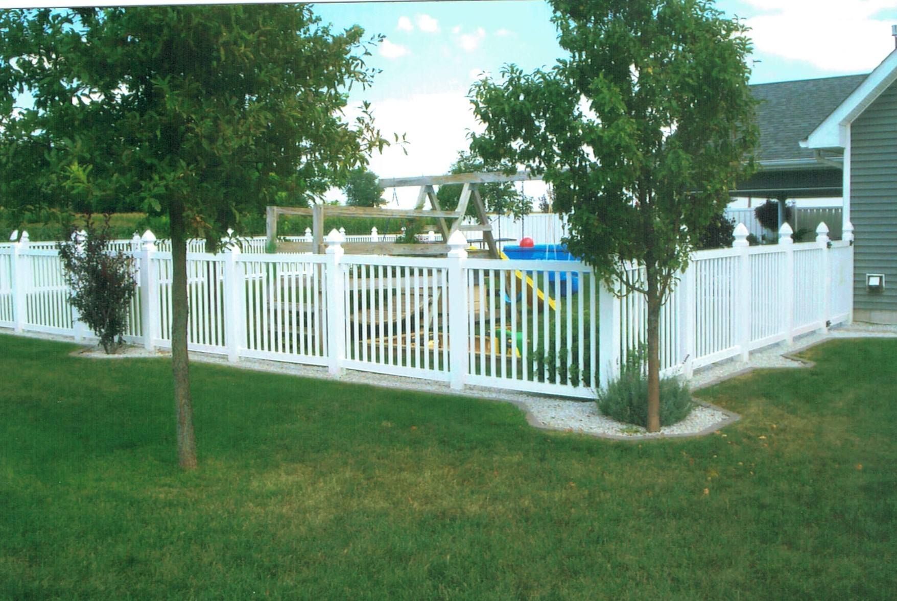 A white picket fence encloses a backyard playground with a small slide, surrounded by green grass and two young trees.
