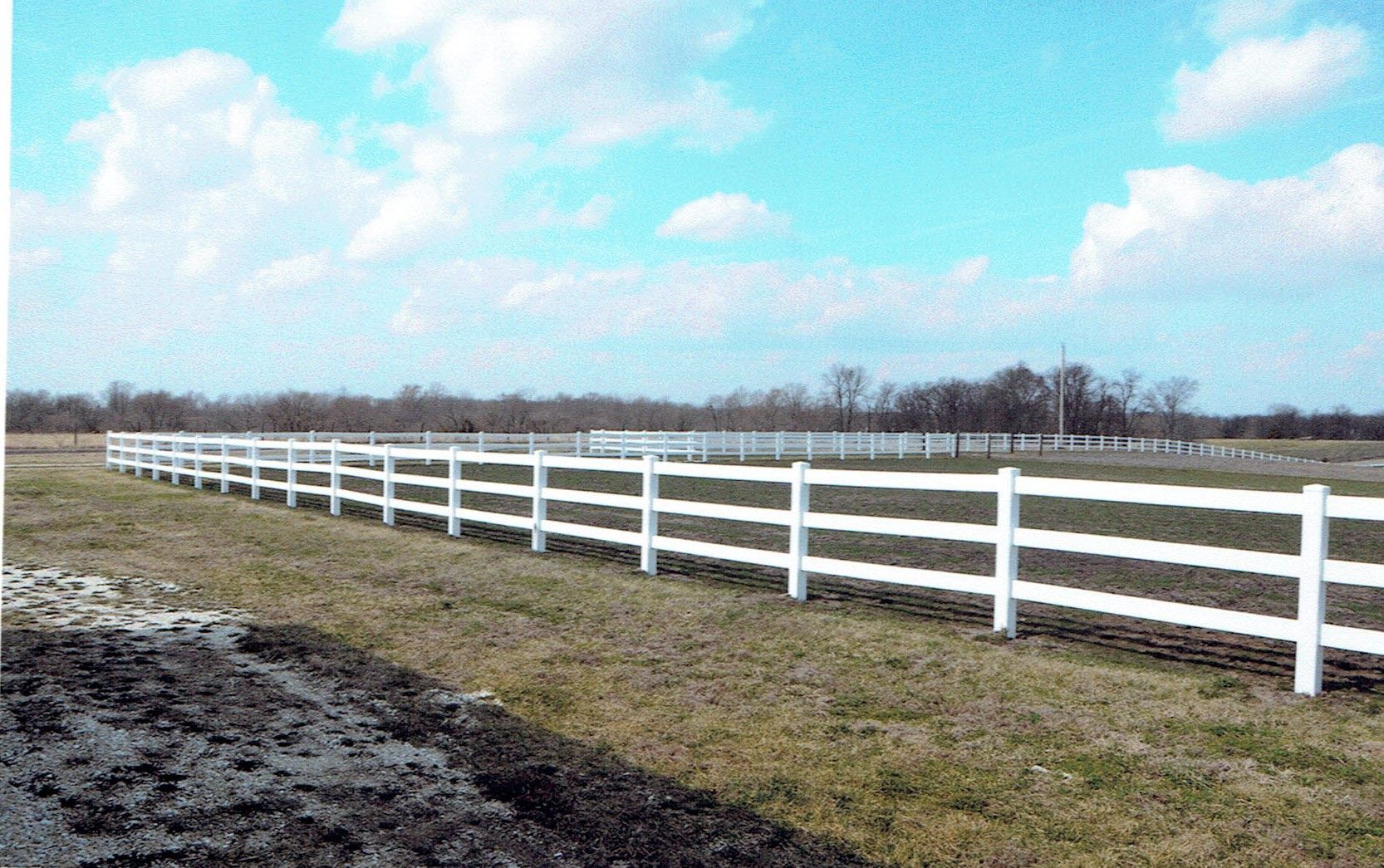 A white three-rail fence curves through an open field under a blue sky with light clouds.