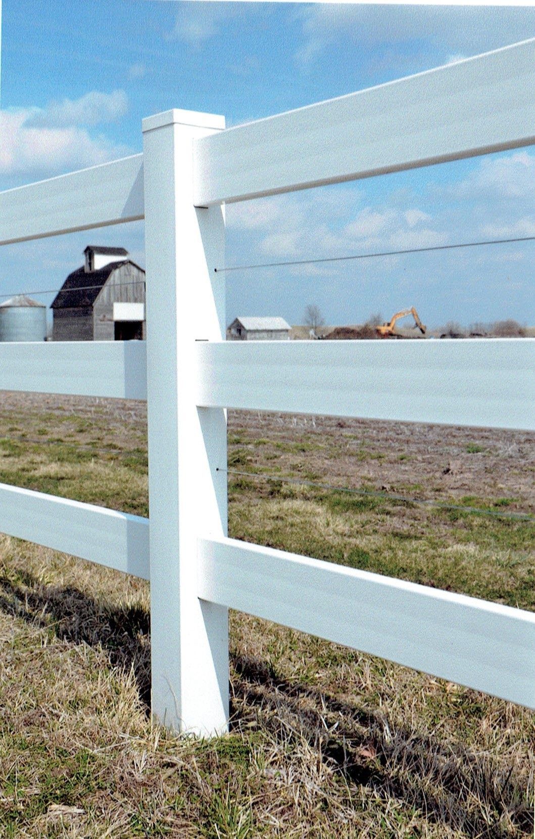 A white, three-rail vinyl fence post stands in a grassy field with a barn and construction equipment in the background.