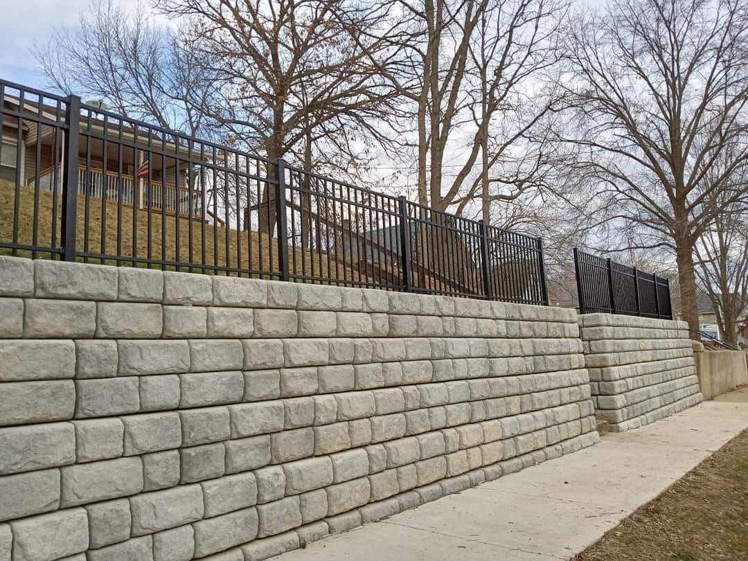 A tall gray stone retaining wall topped with a black metal fence beside a paved sidewalk under bare trees.