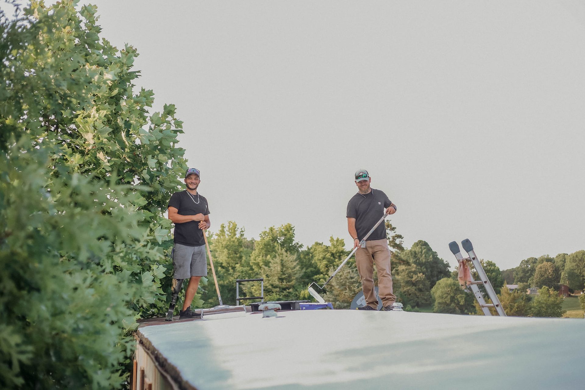 Two men on a roof, holding tools. One smiles, the other looks down. Ladders and trees are visible.